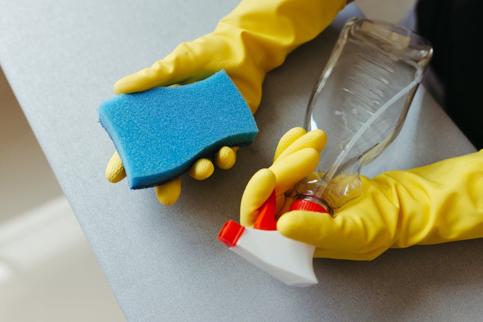 Close-up of a person wearing yellow rubber cleaning gloves holding a blue cleaning sponge and a spray bottle with a clear plastic container, which contains a cleaning solution. The sponge is slightly worn, and the spray bottle has a red trigger. The scene takes place on a light grey countertop, likely in a domestic kitchen or bathroom, with clean, smooth surfaces and bright lighting highlighting the freshness and cleanliness. [COMPANY_NAME] specializes in surface cleaning and sanitisation services, ensuring hygienic and spotless environments. The background is softly blurred, emphasizing the cleaning tools and process, aligned with deep cleaning and routine maintenance for hygiene and surface care.