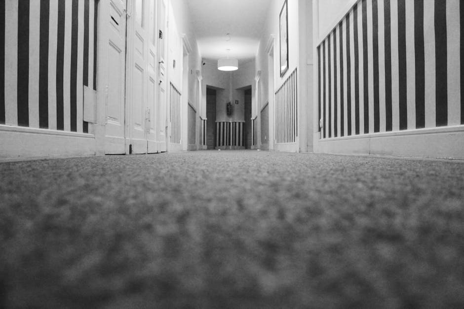 A low-angle black and white photograph of a corridor in a residential or commercial building, showing a textured carpeted floor extending towards the end of the hall. The corridor is lined with white panelled walls and doors on each side, with vertical striped wallpaper running above the panels. a ceiling-mounted circular light fixture provides illumination. The cleanliness of the carpet suggests recent or professional deep cleaning, consistent with surface cleaning and sanitisation services offered by Carpet Cleaning Sidcup to maintain hygiene in indoor environments.